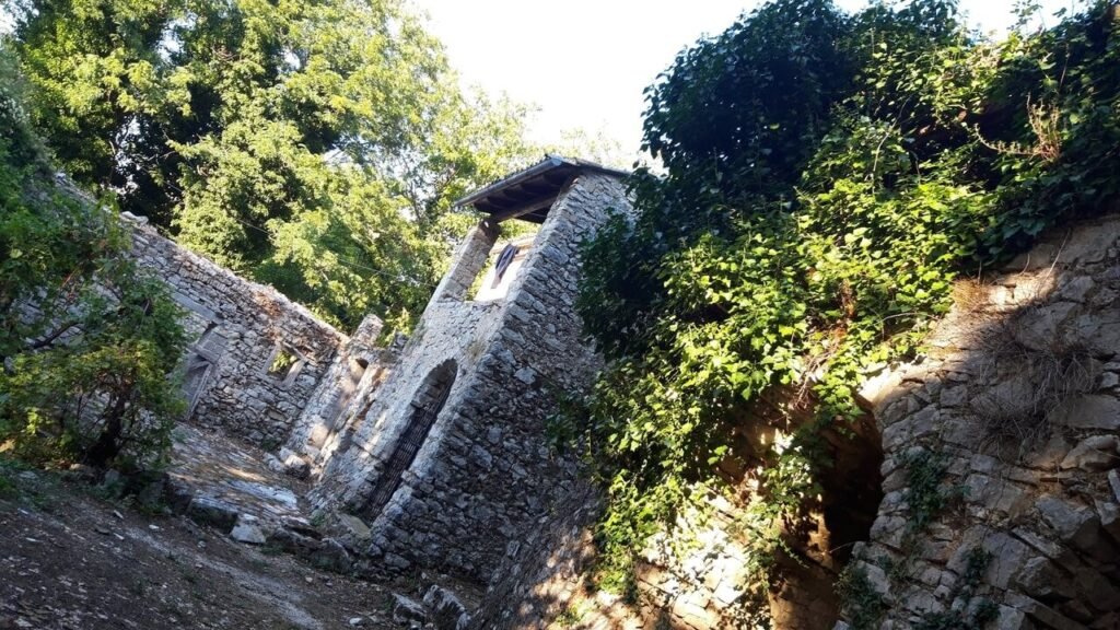 Κοντινή λήψη από χαμηλή γωνία των ερειπωμένων πέτρινων τοίχων ενός κτιρίου. Η κατασκευή είναι φτιαγμένη από μεγάλες, γκρι πέτρες και καλύπτεται μερικώς από πυκνή, πράσινη κισσόφυτη βλάστηση. Στη δεξιά πλευρά διακρίνεται μια πέτρινη καμάρα, ενώ πιο ψηλά στέκει ένα σκεπαστό τμήμα με άνοιγμα. Η συνολική εικόνα αποπνέει την αίσθηση ενός ιστορικού, εγκαταλελειμμένου μνημείου, όπου η πέτρα και το πράσινο συνθέτουν ένα σκηνικό φθοράς και παλαιότητας.