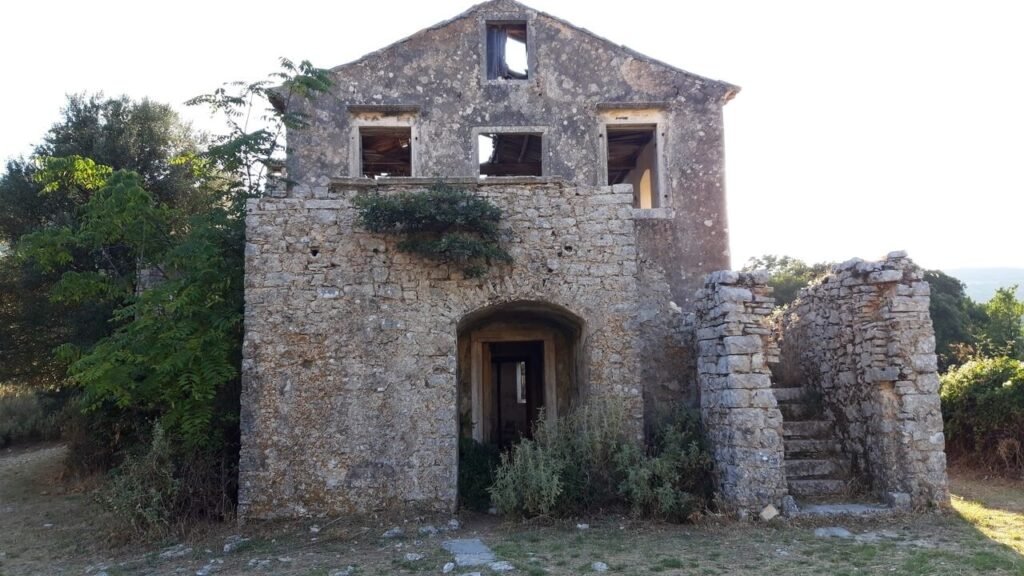 Εγκαταλελειμμένο πέτρινο σπίτι στην Παλιά Περίθεια της Κέρκυρας, με φθαρμένους τοίχους, άδεια πλαίσια παραθύρων και κατεστραμμένη σκεπή. Το τοξωτό άνοιγμα της εισόδου οδηγεί στο ερειπωμένο εσωτερικό, ενώ κισσός και θάμνοι αναπτύσσονται γύρω από το κτίσμα. Δεξιά, μια κατεστραμμένη πέτρινη σκάλα θυμίζει την παλιά ζωή του χωριού, ανάμεσα στο πράσινο και το απαλό απογευματινό φως.
