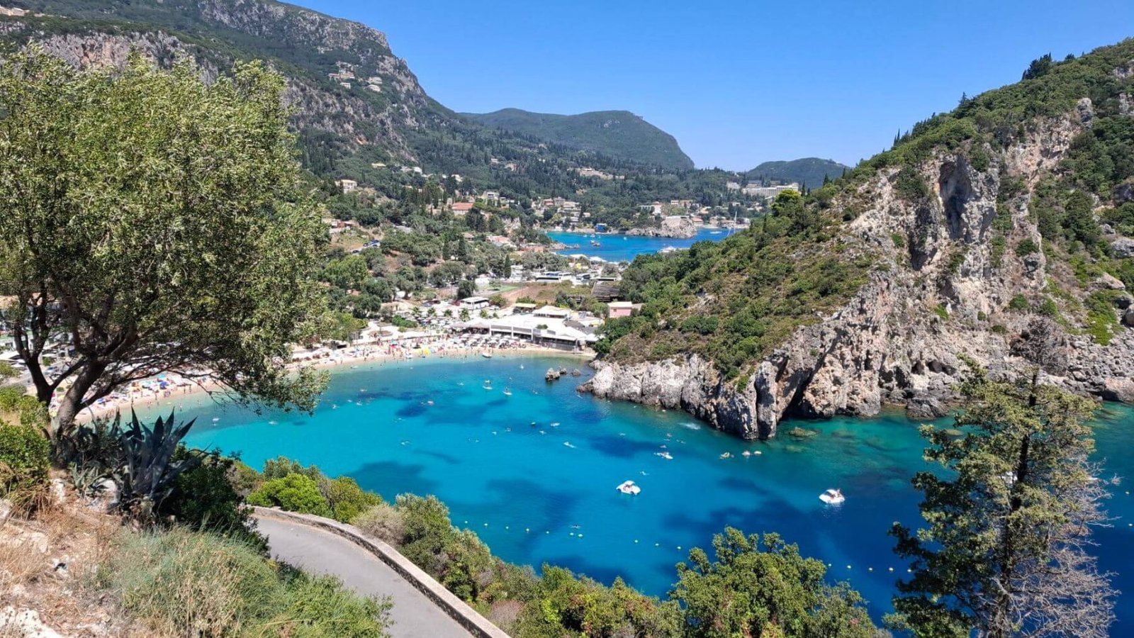 A scenic view of a turquoise bay surrounded by lush green hills and rocky cliffs, with boats on the water, a beach, and a village in the background under a clear blue sky.